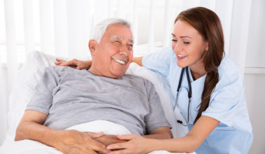 Nurse Looking At Happy Male Patient Lying On Bed In Clinic