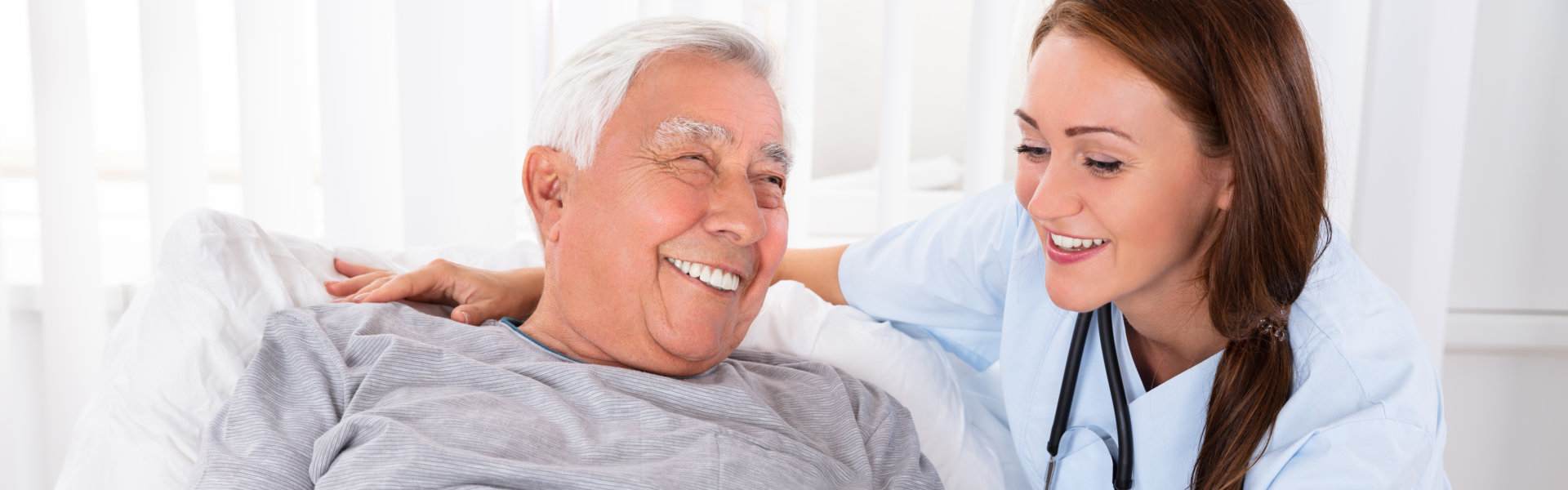 Nurse Looking At Happy Male Patient Lying On Bed In Clinic